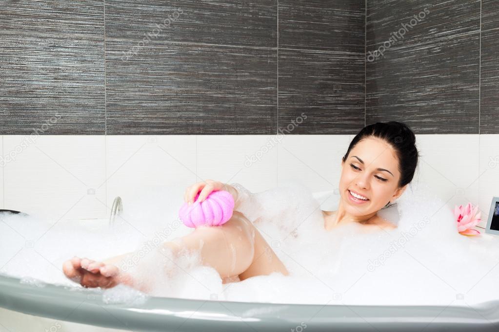 Beautiful smile woman washing leg with pink sponge in bath with foam