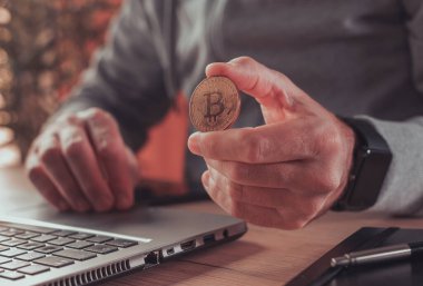 Girişimci Bitcoin kripto para ile ticaret yapıyor. Dizüstü bilgisayarın önünde altın sikke tutan bir erkeğin kavramsal görüntüsü.