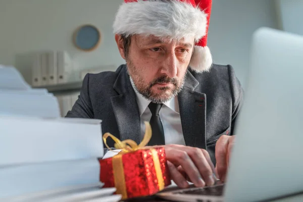 彼のオフィスでクリスマス休暇シーズン中にノートパソコンのキーボードを入力するサンタクロースの帽子を持つ深刻なビジネスマン 選択的な焦点 — ストック写真