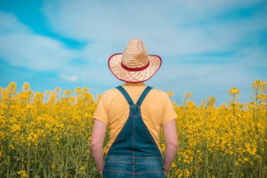 Dikiz aynasından bakan dişi çiftçi tarımcı çiçek açan kolza tarlasında duruyor, ekinlere ve ufka bakıyor.