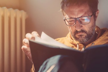 Düşünceli, orta yaşlı bir erkek evde kitap okuyor. Yakın plan, seçici odaklı bir fotoğraf. Pencereden yansıyan güneş ışığı.