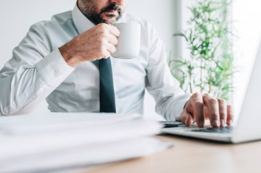 Drinking coffee at work increases productivity, businessman with cup of hot beverage working on laptop computer, selective focus