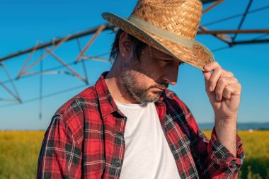 Ciddi bir çiftçinin portresi kolza tohumu tarlasında işlevsiz merkez pivot sulama ekipmanının önünde duruyor, seçici odaklanma