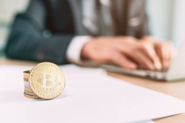 Bitcoin kripto para tüccarı kavramı, ofis masasına istiflenmiş paraların yakın plan görüntüsü ve arka planda dizüstü bilgisayar kullanan iş adamı
