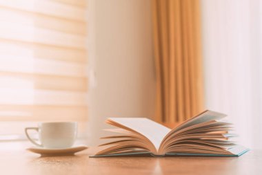 Açık kitap ve kahve fincanı masada, sabahları evde okuyor, seçici bir şekilde odaklanıyor.
