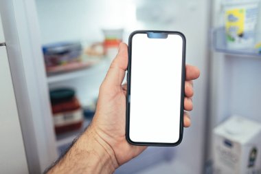 Akıllı telefon modelleme konsepti, buzdolabının önünde boş ekranlı akıllı cep telefonu tutan ve market malzemelerini kontrol eden, seçici odaklı adam.