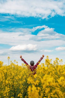 Başarılı bir kadın çiftçi, ekose gömlek ve kollarıyla havaya kaldırılmış kamyoncu şapkası takıyor. Muzaffer bir poz veriyor, çiçek açan kolza tarlasında duruyor ve ufka bakıyor, seçici odaklanıyor.