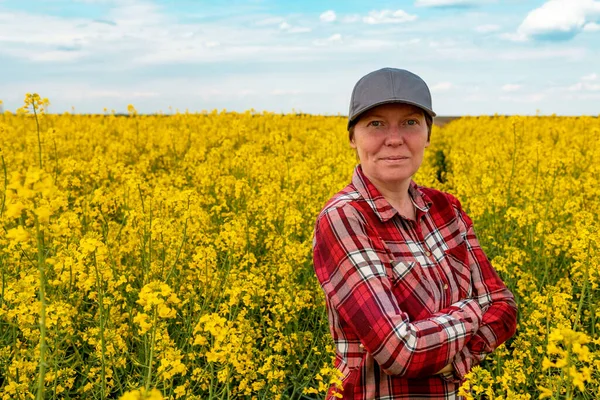 Confident and self-assured farm worker wearing red plaid shirt and trucker\'s hat standing in cultivated rapeseed field in bloom and looking over crops, selective focus