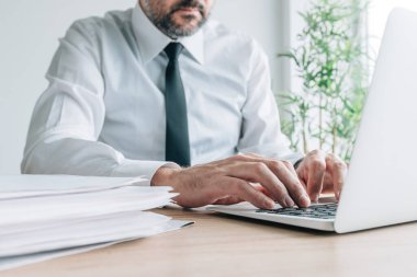 Businessman working on laptop computer with paperwork pile on office desk, selective focus