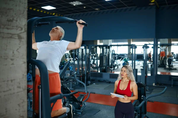 Old man on exercise machine, trainer with laptop — Stock Photo, Image