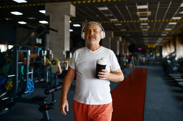Homem velho em fones de ouvido bebe água no ginásio — Fotografia de Stock