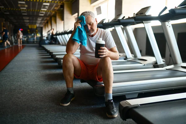 Втомлений старий з рушником п'є воду в спортзалі — стокове фото