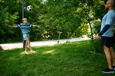 Baba ve çocuk parkta çimlerde futbol oynuyorlar.