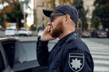 Polis memuru telefonda konuşuyor, araba parkında.