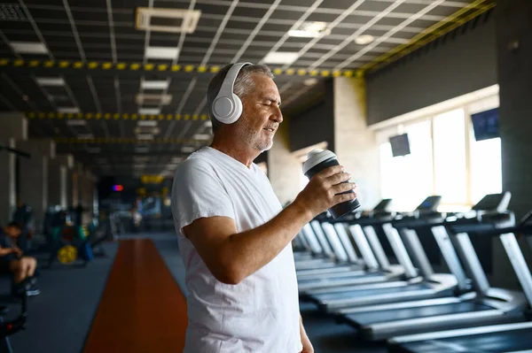 Homem velho em fones de ouvido bebe água no ginásio — Fotografia de Stock