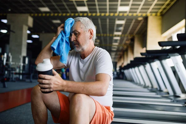 Zmęczony staruszek z ręcznikiem pije wodę na siłowni — Zdjęcie stockowe
