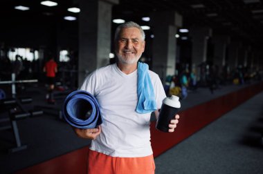 Beden dersinde bir şişe su, havlu ve paspas taşıyan yaşlı bir adam.