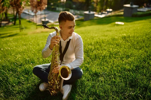 Jovem saxofonista com saxofone sentado na grama — Fotografia de Stock