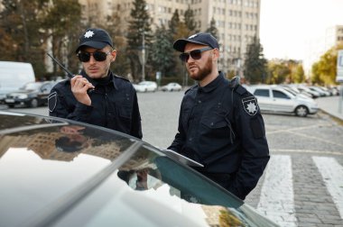 İki erkek polis telsizle arabayı kontrol ediyor.