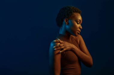 Genç bir kadın koyu arkaplanda omuzlarına dokunuyor. Mavi ışık, stüdyo çekimi.