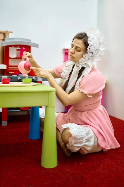 家や保育所でカラフルなお茶セットのおもちゃでお茶パーティーを遊んで赤ちゃんのように身に着けている面白い女性 — ストック写真