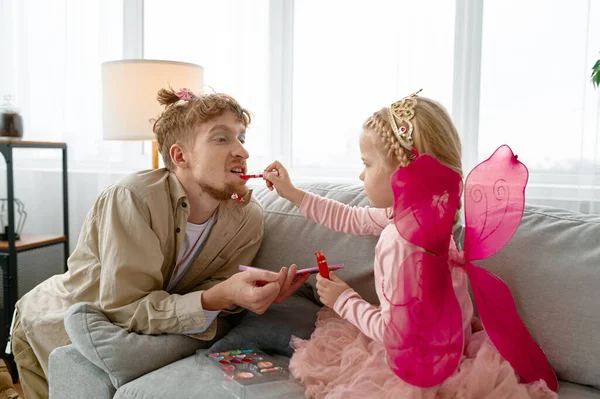 Little daughter putting lipstick on dads lips applying make-up cosmetics enjoy time with parent at home