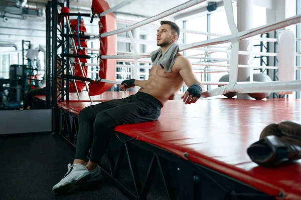 Portrait of muscular male boxer sitting on boxing ring mat and rest after hard training session or competitive match