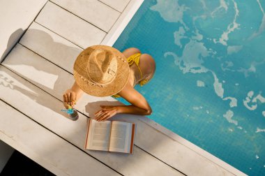Yukarıdan kadınların yüzme havuzunda okuduğu ve içtiği ferahlatıcı kokteyli izleyin. Tatil ve seyahat kavramı
