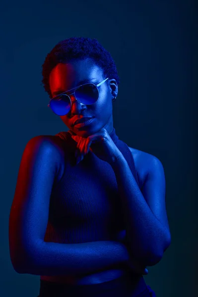 Pensive woman in sunglasses over dark studio background with copy space and neon light