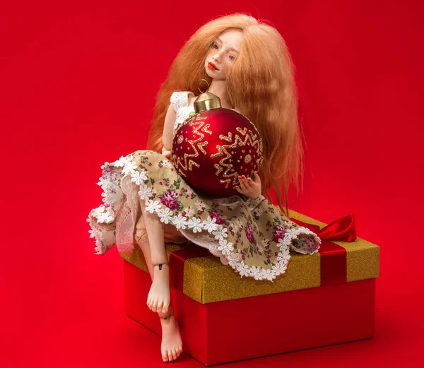 Vánoční Míč Rukou Krásné Panenky Sedí Krabici Dárkem Červeném Pozadí — Stock fotografie