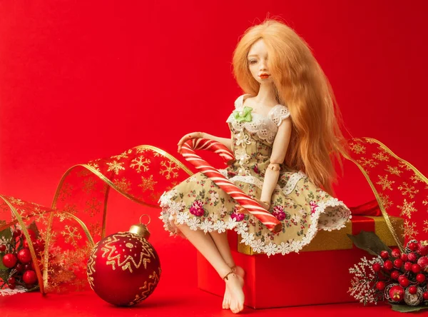 一个漂亮的洋娃娃手里拿着焦糖手杖坐在盒子上 盒子里有红色背景的礼物和圣诞装饰品 — 图库照片
