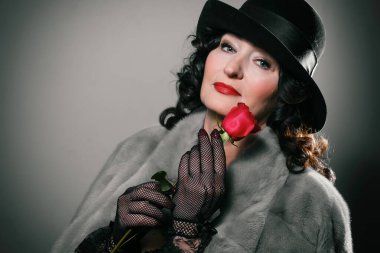 Kürk ceketli, şapkalı ve eldivenli güzel bir kadın ellerinde dudaklarında kırmızı bir gül tutuyor.