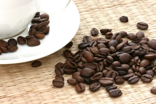 Celá kávová zrna roztroušených na slaměné pozadí s bílou cup — Stock fotografie