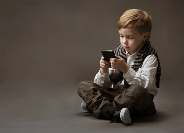 Chico jugando en el teléfono celular. Niño sentado sobre fondo gris a —  Fotos de Stock
