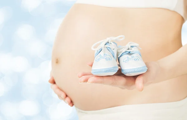 Barriga de mulher grávida com botas de bebê recém-nascido close-up sobre blu — Fotografia de Stock