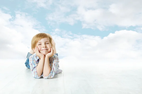 Улыбающийся ребенок лежит, маленький ребенок смотрит в камеру. Синее небо и облака — стоковое фото