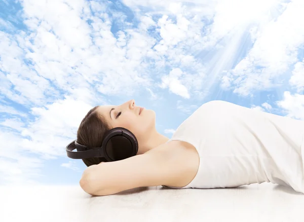 Woman in headphones dreaming listening to music. Girl relaxing over blue sky background — Stock Photo, Image