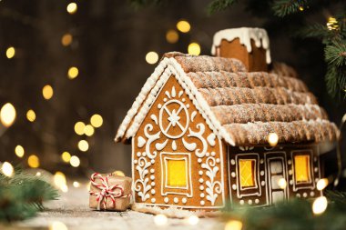 Gingerbread House with Christmas Gift over Shining Garland Lights Background. Xmas Holiday Present Art Decoration. Baked Sweet Ginger Cake with Icing clipart