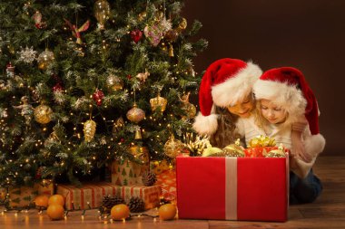 Büyük kırmızı hediye kutusu hediye oyuncaklarıyla dolu Noel çocukları. Xmas Ağacı 'nın yanındaki iki küçük kız Kahverengi Oda Arkaplanının üzerindeki altın süslemelerle süslenmiş.