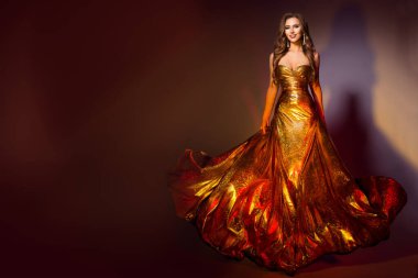 Uzun Altın Elbiseli Moda Modeli. Gece vakti gülümseyen güzel kadın Koyu kahverengi arka planda yürüyen kanat çırpan altın elbise