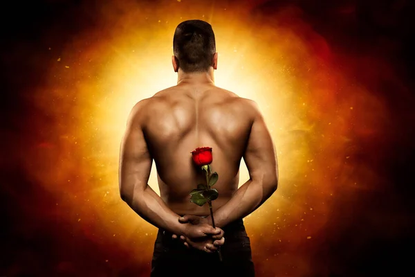 男のリアビュー隠された花の贈り物神秘的な輝く光に対して後ろに戻る 芸術的なファンタジーの背景に赤いバラと強力な筋肉のトップレスボディビルダーの背景 — ストック写真