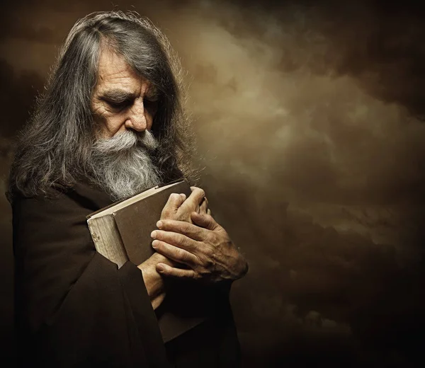 İncil 'le dua eden keşiş. Peygamber kitabı tutuyor. Uzun Gri Sakallı Yaşlı Adam portresi Kara Gizemli Arkaplan Üzerinde Siyah Pelerinli