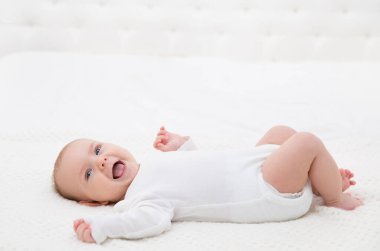Mutlu Gülümseyen Bebek Beyaz Yatak 'ta uzanıyor. Beyaz Pamuklu Giysili Aktif Bebek Çocuk. Bebeklerin içinde gülen bebek bebek tulumları kameraya bakıyor. Evde Üç Ay Küçük Çocuk