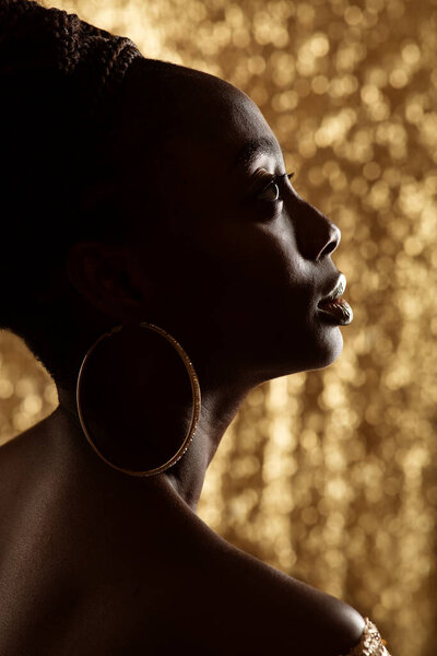 African Woman Face Profile Black Silhouette with Large Golden Rings Earrings. Dark Skinned Beauty Model with Gold Jewelry over shining Bokeh Background looking up