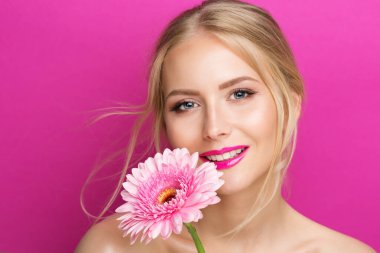 Pembe rujlu güzellik modeli. Yüzü pürüzsüz cilt makyajı ve dudak parlatıcısı ile gülümseyen genç kadın. Mor Arkaplanda Çiçekli Parlak Rujlu Moda Kızı Portresi. Kadınlar Kozmetik ve Yaz Yüz Bakımı