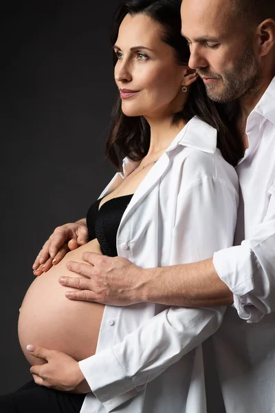 Pregnant Couple in White Shirts embracing Belly over Black Background. Mother and Father Love expecting Baby during Pregnancy. Family waiting Child Birth. Man Hands on Pregnant Woman naked Abdomen