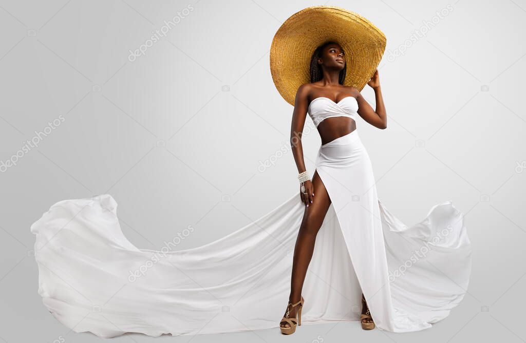 Fashion Model in White Dress and Big Summer Hat. Sexy Stylish Woman in Long Slit Gown flying on Wind showing tanned Leg over isolated Studio Background