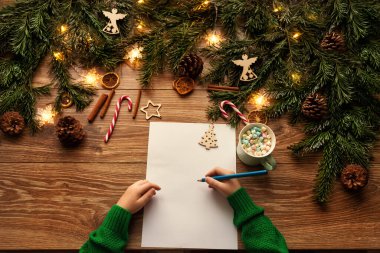 Noel Baba 'ya mektup. Noel Çocuk Elleri Kahverengi Tahta Masa 'nın üzerine Noel Hediyeleri Alışveriş Dilekleri Listesi' ni yazıyor. Ağaç dallı ahşap zemin süslenmiş Noel Işıkları ve Oyuncakları