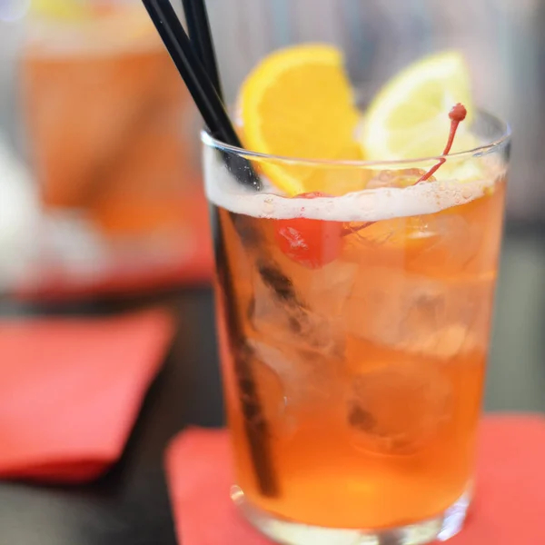 Iced bright orange cocktail with black straws, slice of orange and lemon, maraschino cherry and ice cubes in transparent glass on red napkin. Very selective focus