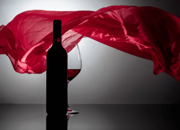 在黑色的反光背景上 盛满了红酒的瓶子和酒杯 背景上红布飘扬 — 图库照片